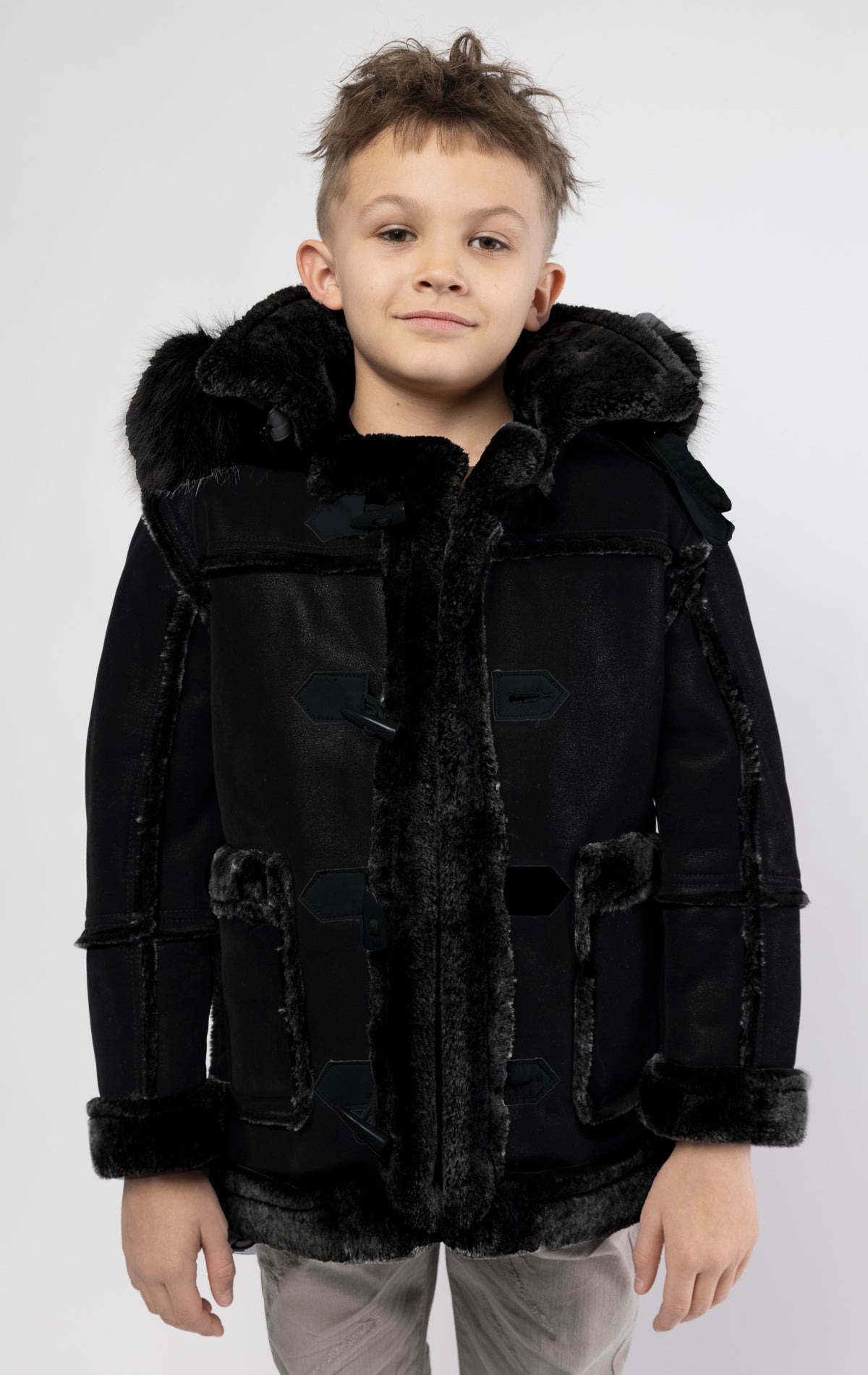 Black jacket lined with plush faux shearling throughout the body and sleeves. It also features shearling-accented pockets, a horn toggle front closure, and vegan suede straps with a buckle at the neck. For added convenience, a detachable faux fox fur hood with a zipper is included. This product is also available for adults.