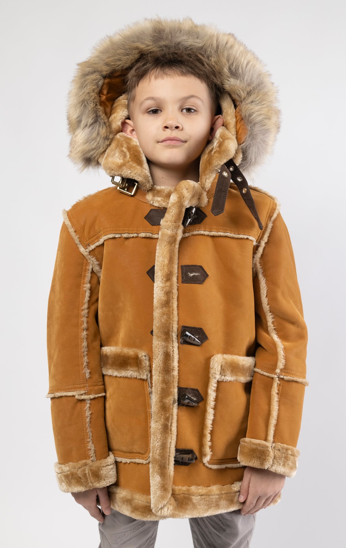 Cognac jacket lined with plush faux shearling throughout the body and sleeves. It also features shearling-accented pockets, a horn toggle front closure, and vegan suede straps with a buckle at the neck. For added convenience, a detachable faux fox fur hood with a zipper is included. This product is also available for adults.