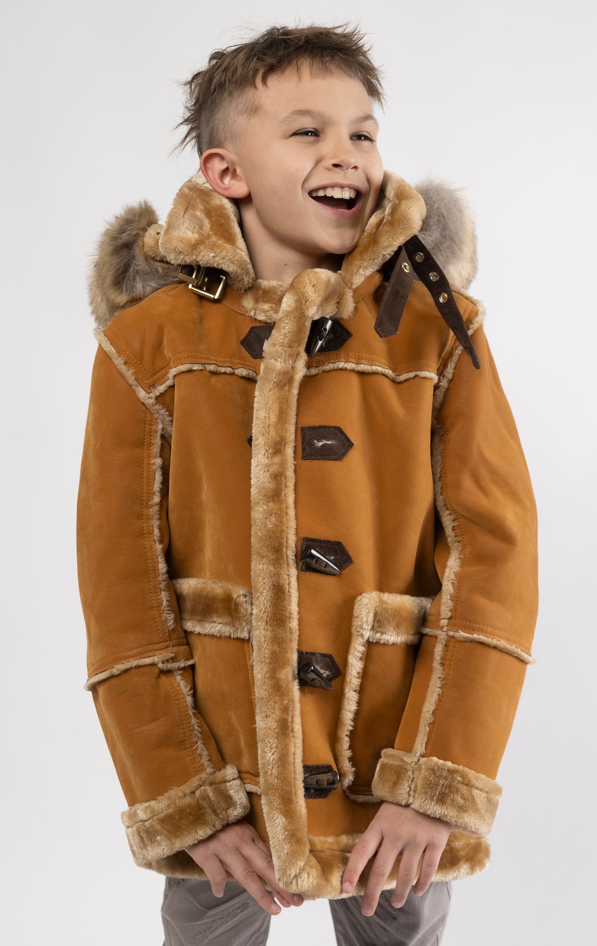 Cognac jacket lined with plush faux shearling throughout the body and sleeves. It also features shearling-accented pockets, a horn toggle front closure, and vegan suede straps with a buckle at the neck. For added convenience, a detachable faux fox fur hood with a zipper is included. This product is also available for adults.