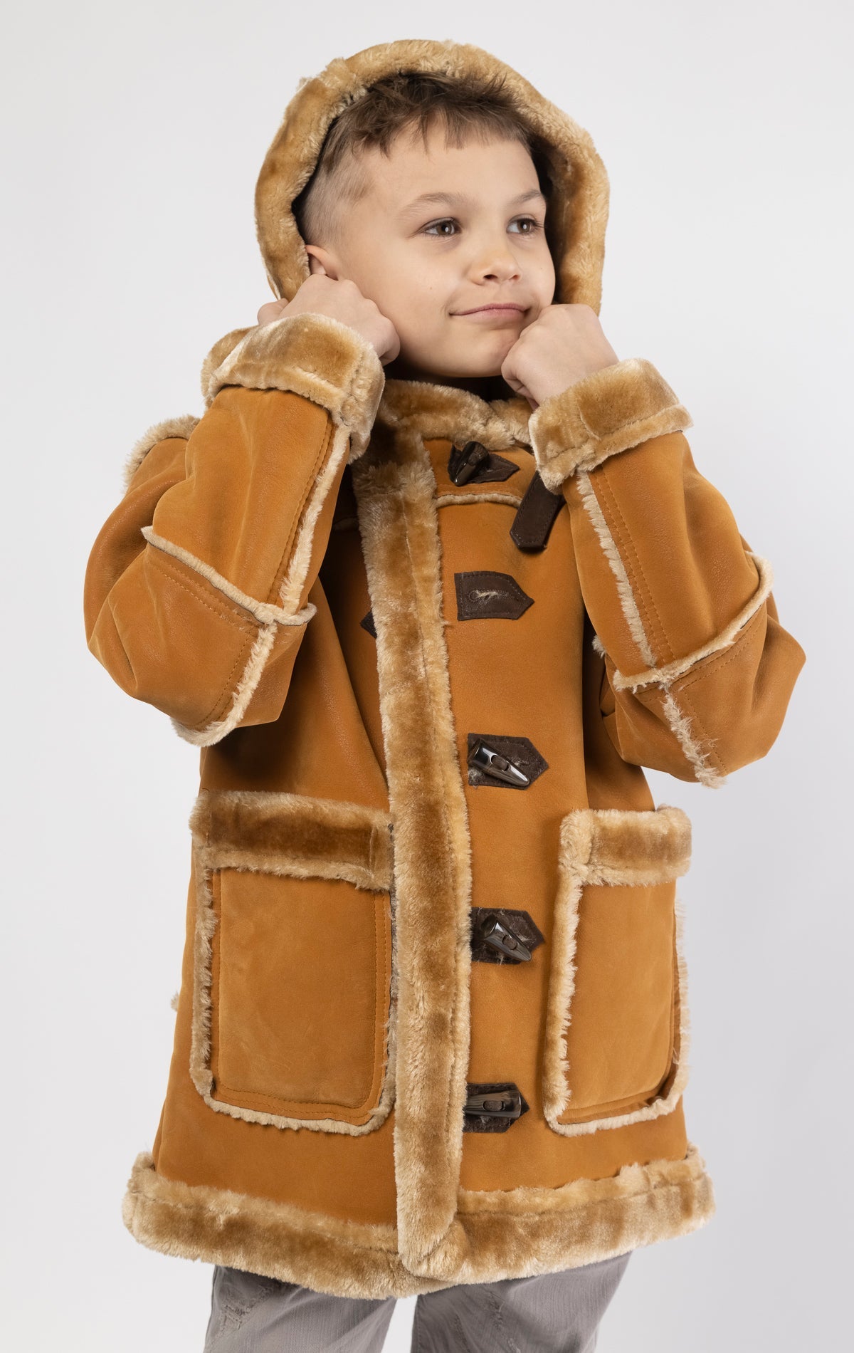 Cognac jacket lined with plush faux shearling throughout the body and sleeves. It also features shearling-accented pockets, a horn toggle front closure, and vegan suede straps with a buckle at the neck. For added convenience, a detachable faux fox fur hood with a zipper is included. This product is also available for adults.