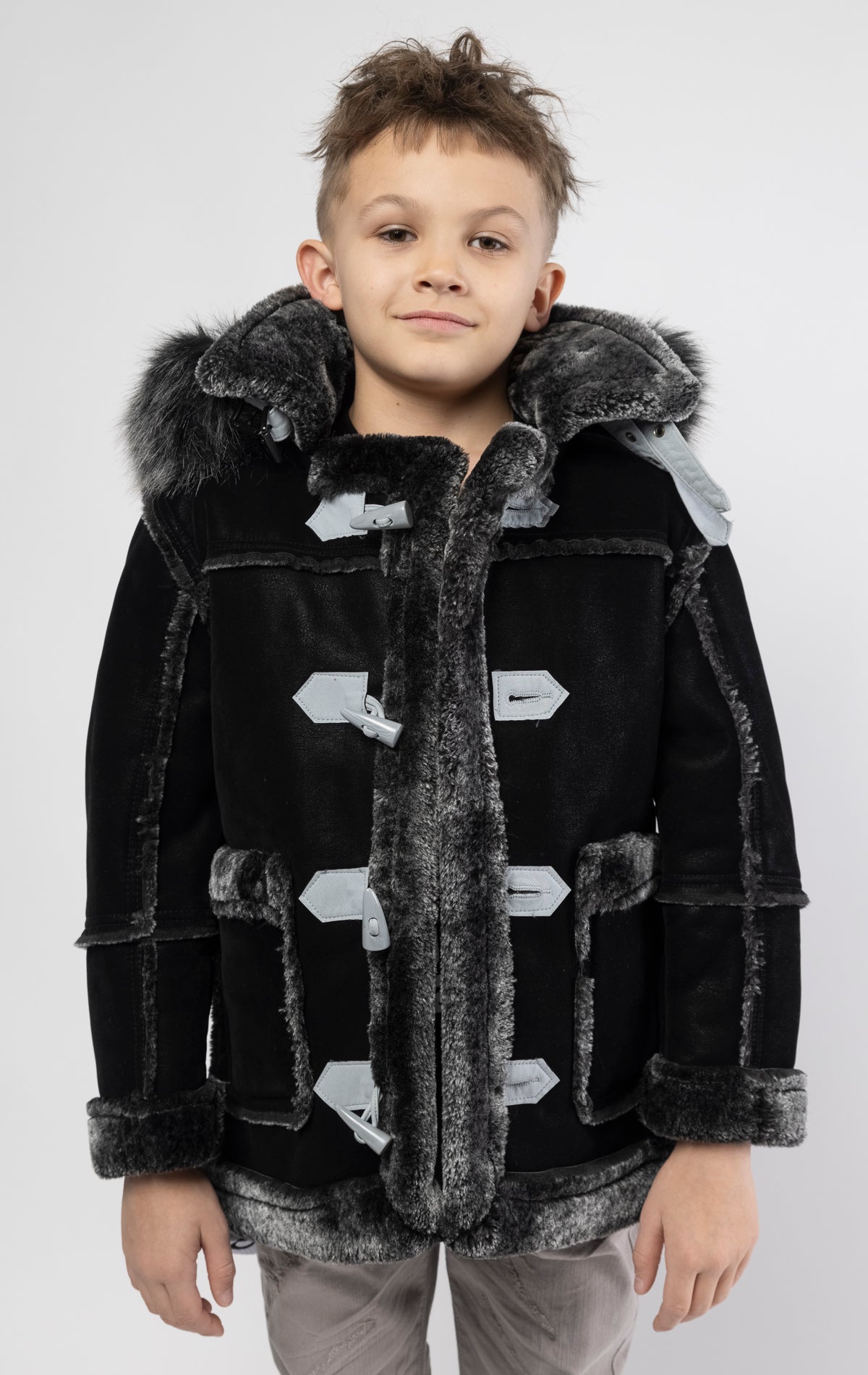 Black jacket lined with plush faux shearling throughout the body and sleeves. It also features shearling-accented pockets, a horn toggle front closure, and vegan suede straps with a buckle at the neck. For added convenience, a detachable faux fox fur hood with a zipper is included. This product is also available for adults.