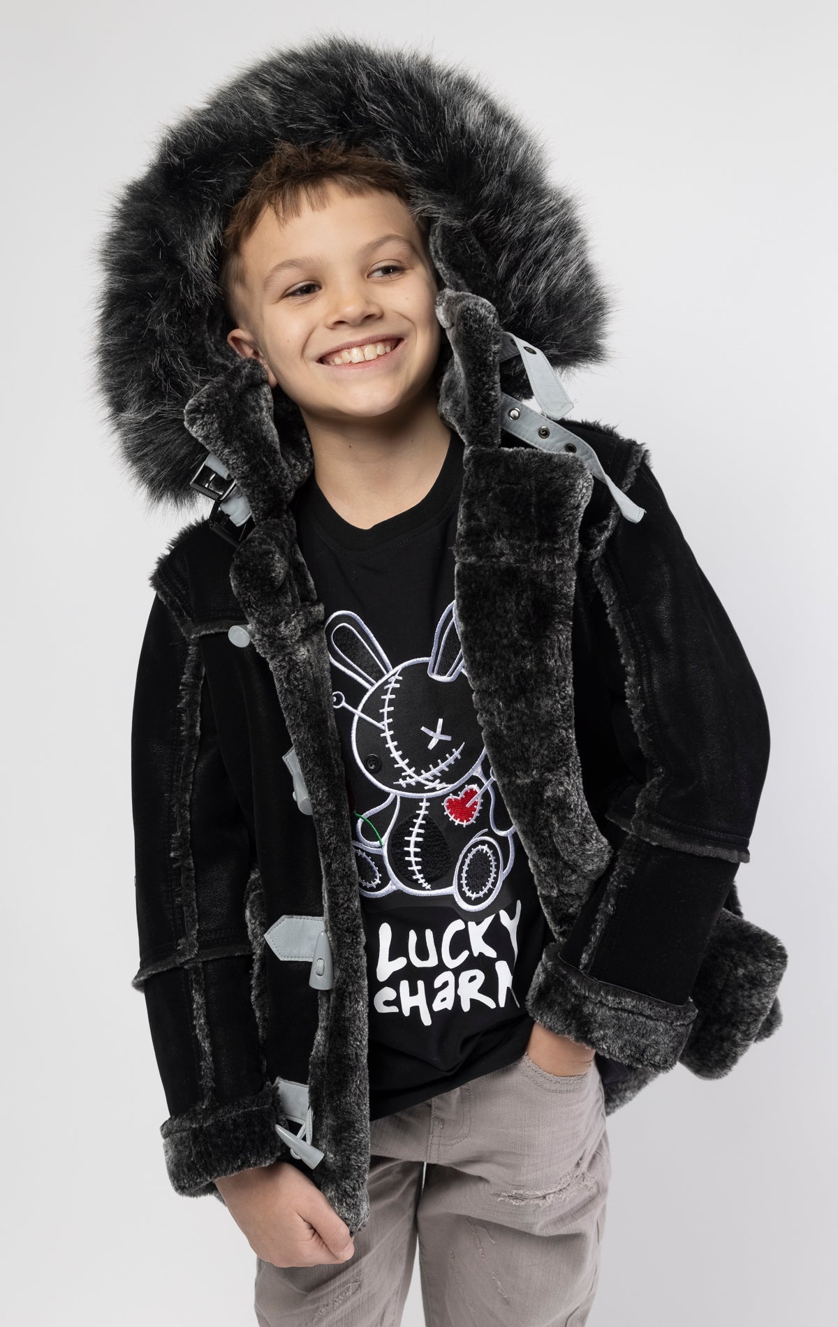 Black jacket lined with plush faux shearling throughout the body and sleeves. It also features shearling-accented pockets, a horn toggle front closure, and vegan suede straps with a buckle at the neck. For added convenience, a detachable faux fox fur hood with a zipper is included. This product is also available for adults.