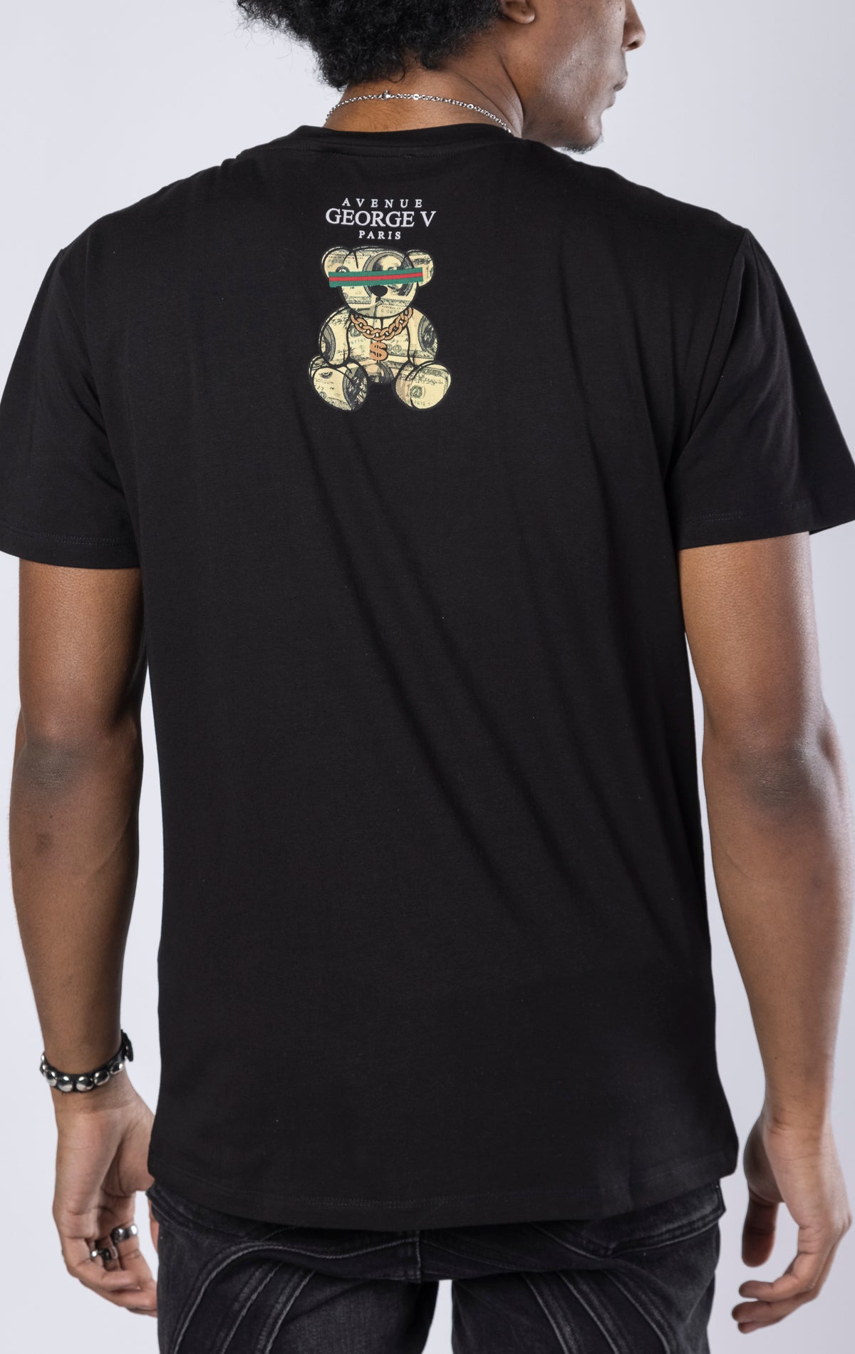 black T-shirt featuring a large, rhinestone-encrusted dollar sign plush on the front. 100% cotton, crewneck, short sleeves.
