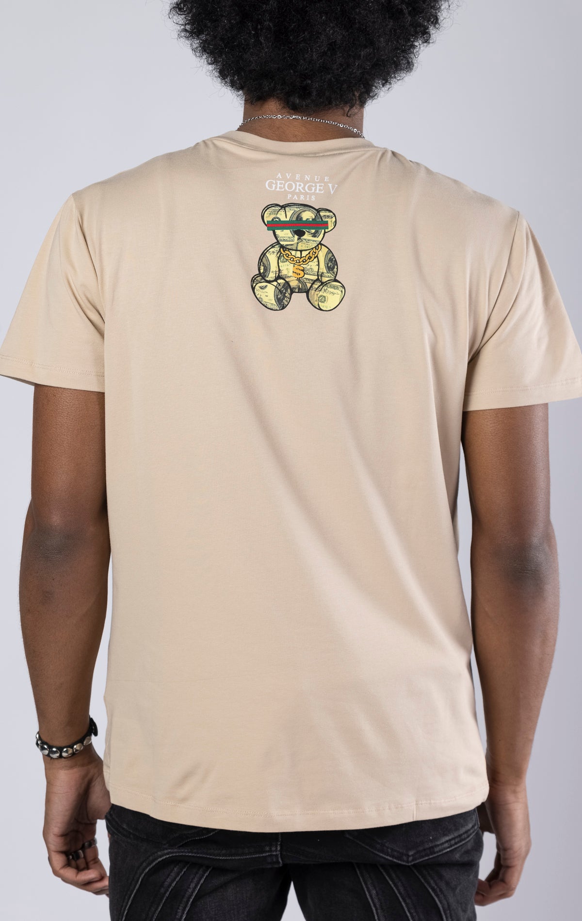 beige T-shirt featuring a large, rhinestone-encrusted dollar sign plush on the front. 100% cotton, crewneck, short sleeves.
