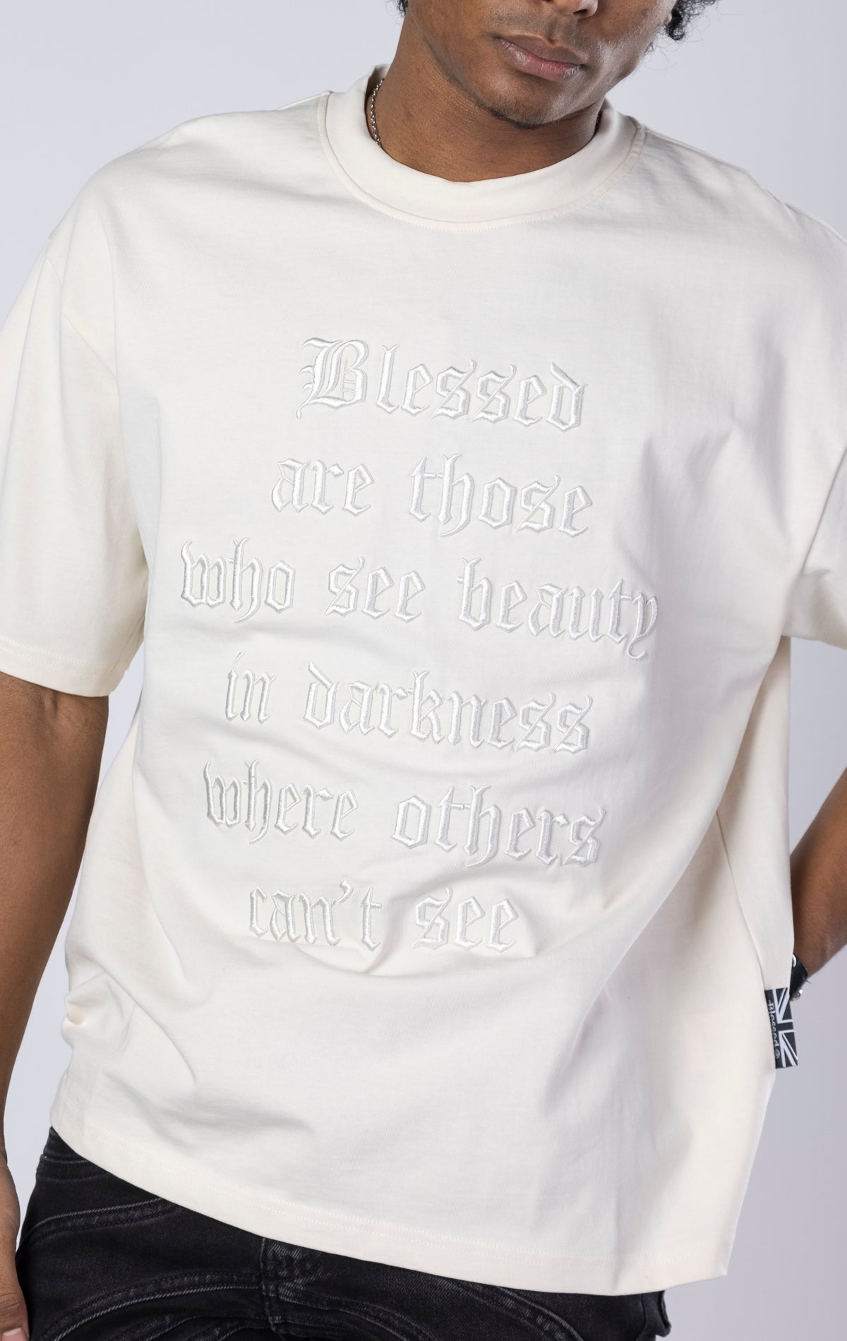 Blessed Darkness Boxy Tee. Cream t-shirt made from 100%  250gr cotton. Features a boxy fit, dropped shoulders, longer sleeves, a 3D embroidered front message, and subtle "blessed" branding