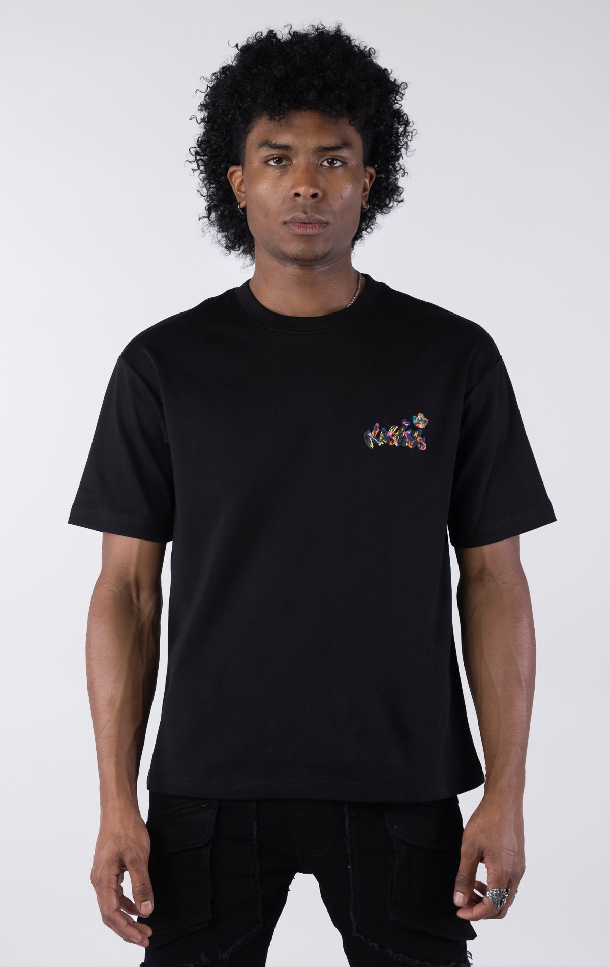 Black short-sleeve t-shirt showing the back, featuring a large, colorful, graffiti-style illustration of a cartoon character in a backwards cap and crown. The text 