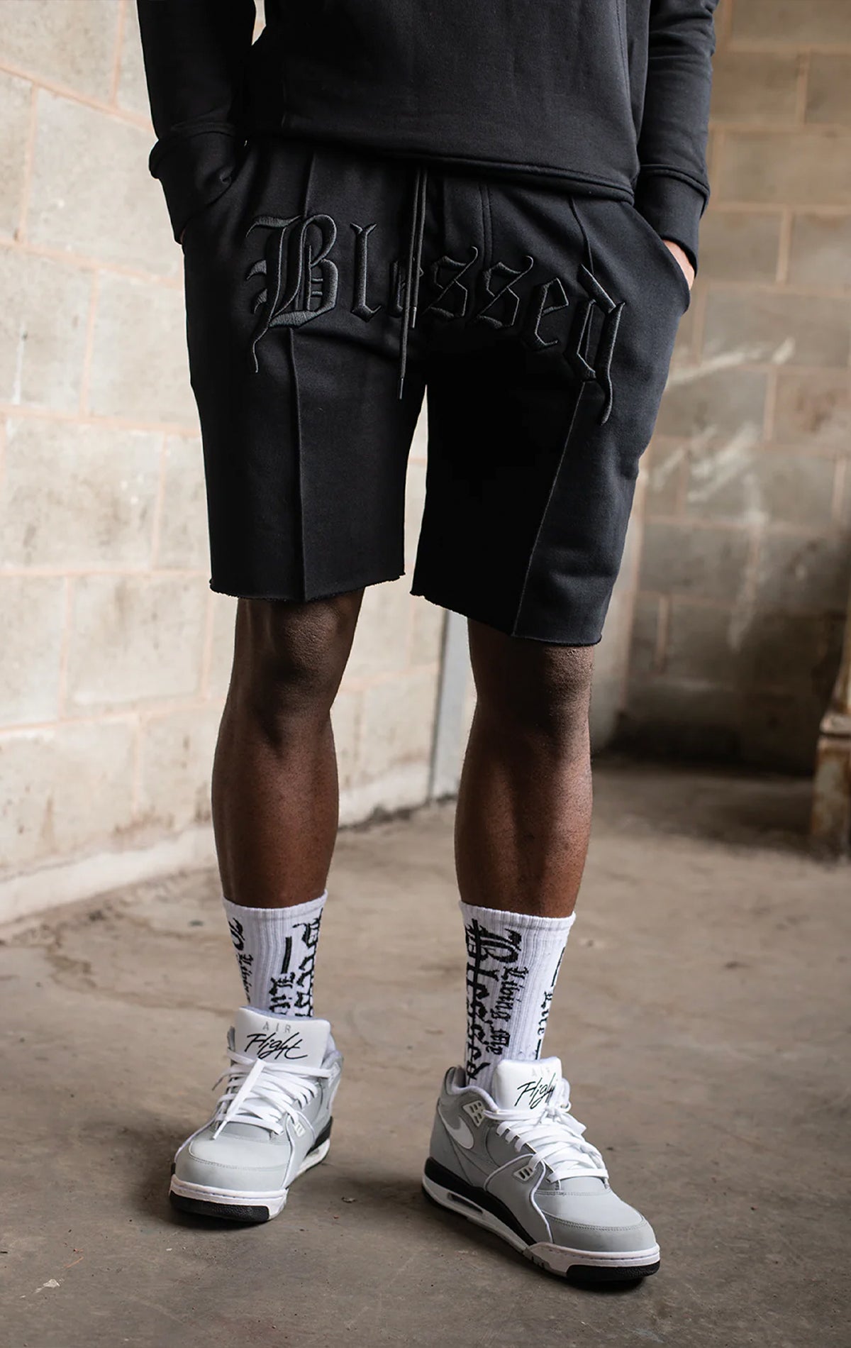 Men's casual, straight-leg shorts in black. The shorts feature a 100% cotton fabric, an arched 3D embroidered "blessed" logo on the front, a tonal raised "blessed" script logo on the back, a back jet pocket, tubular waist drawstrings with metal trims, and a raw hem.
