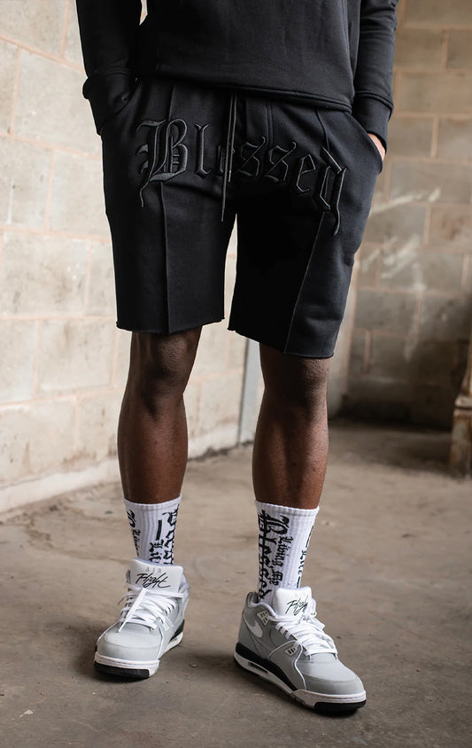 Men's casual, straight-leg shorts in black. The shorts feature a 100% cotton fabric, an arched 3D embroidered "blessed" logo on the front, a tonal raised "blessed" script logo on the back, a back jet pocket, tubular waist drawstrings with metal trims, and a raw hem.
