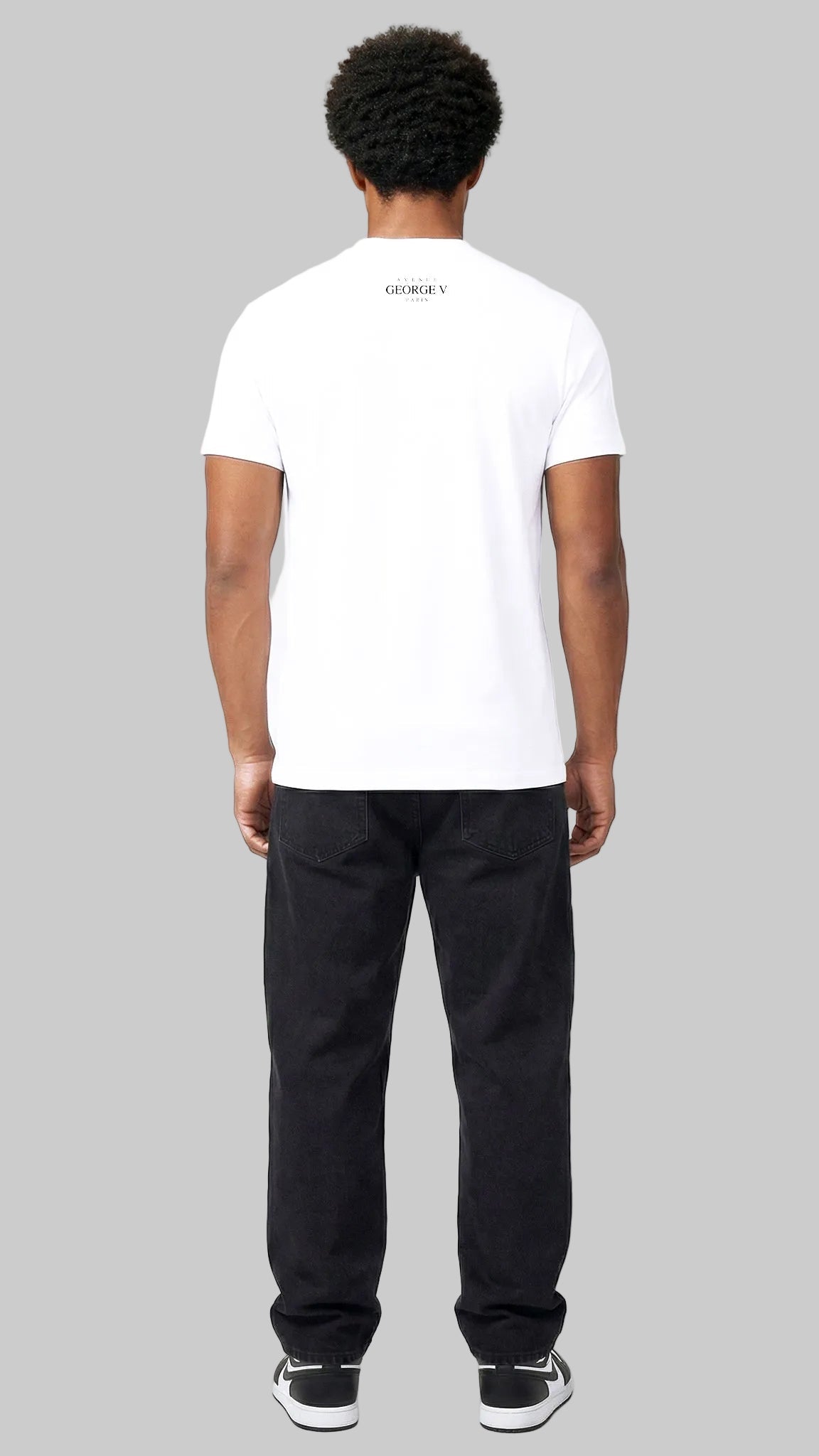 A man viewed from behind wearing a white short-sleeve t-shirt with a small black logo on the upper back, paired with black pants and black-and-white sneakers, against a plain white studio background.