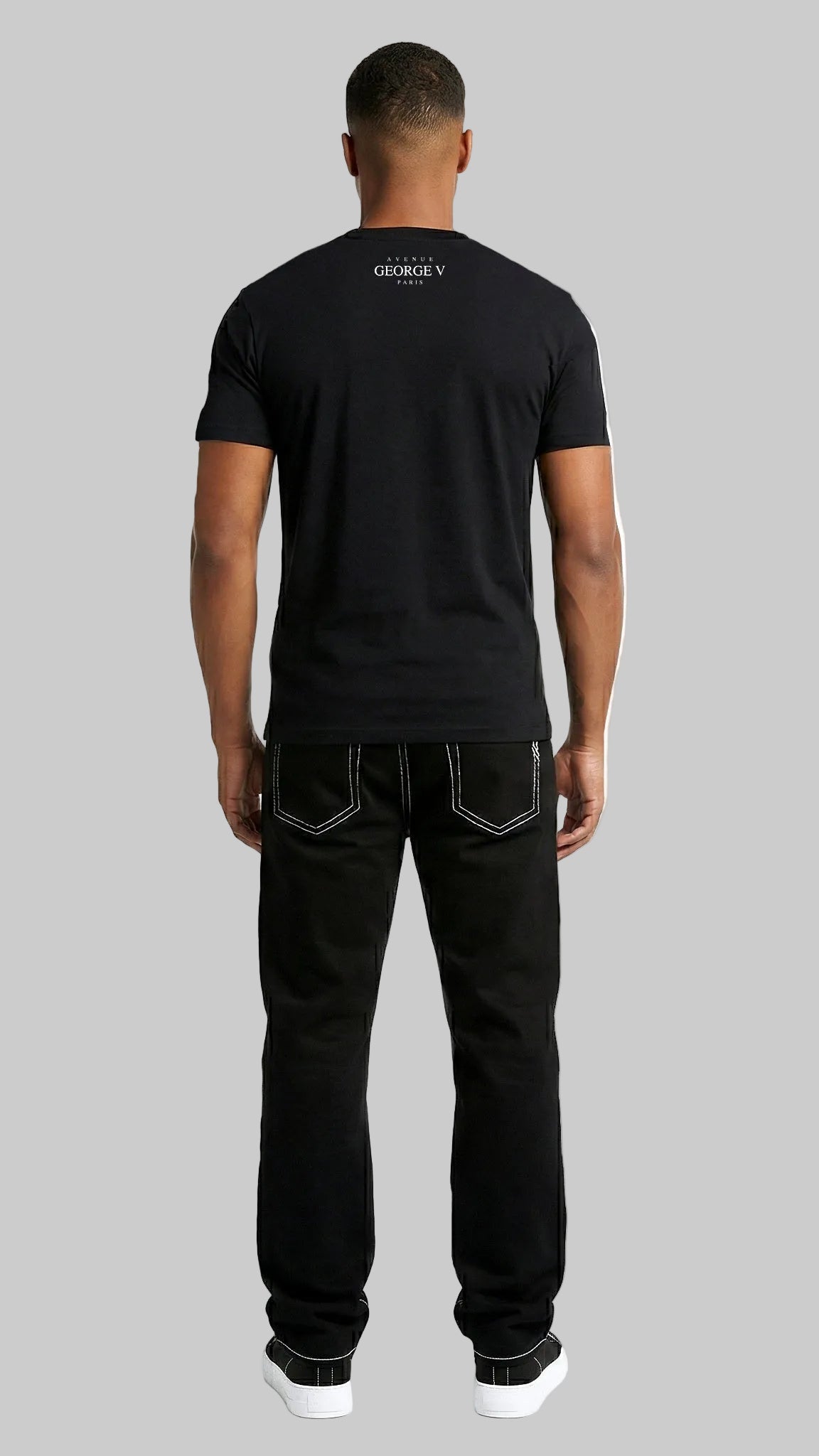 A man viewed from behind wearing a black short-sleeve t-shirt with a small white logo on the upper back, paired with black jeans and white sneakers, against a plain white studio background.