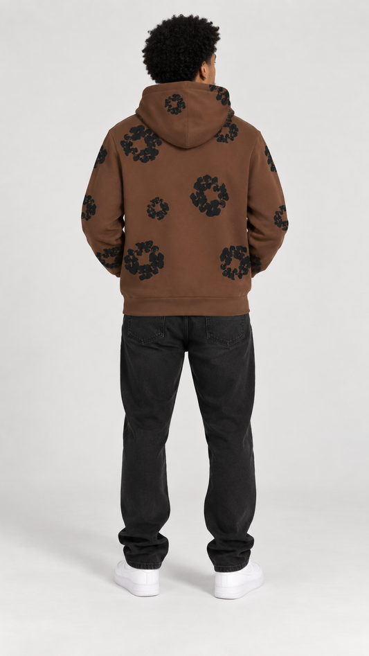 A man viewed from the back wearing a brown pullover hoodie with a black circular graphic pattern, paired with black jeans and white sneakers, against a light gray studio background.