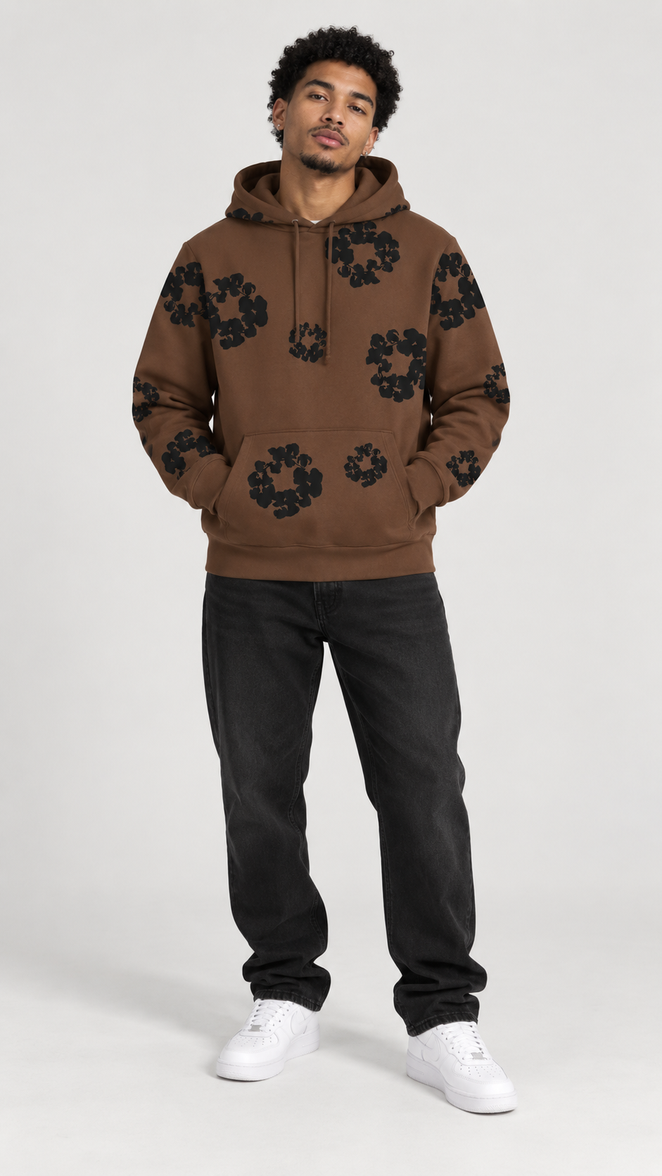 A man modeling a brown pullover hoodie with black floral patterns, paired with black jeans and white sneakers, against a light gray studio background.