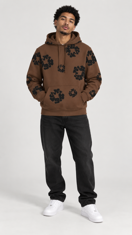 A man modeling a brown pullover hoodie with black floral patterns, paired with black jeans and white sneakers, against a light gray studio background.