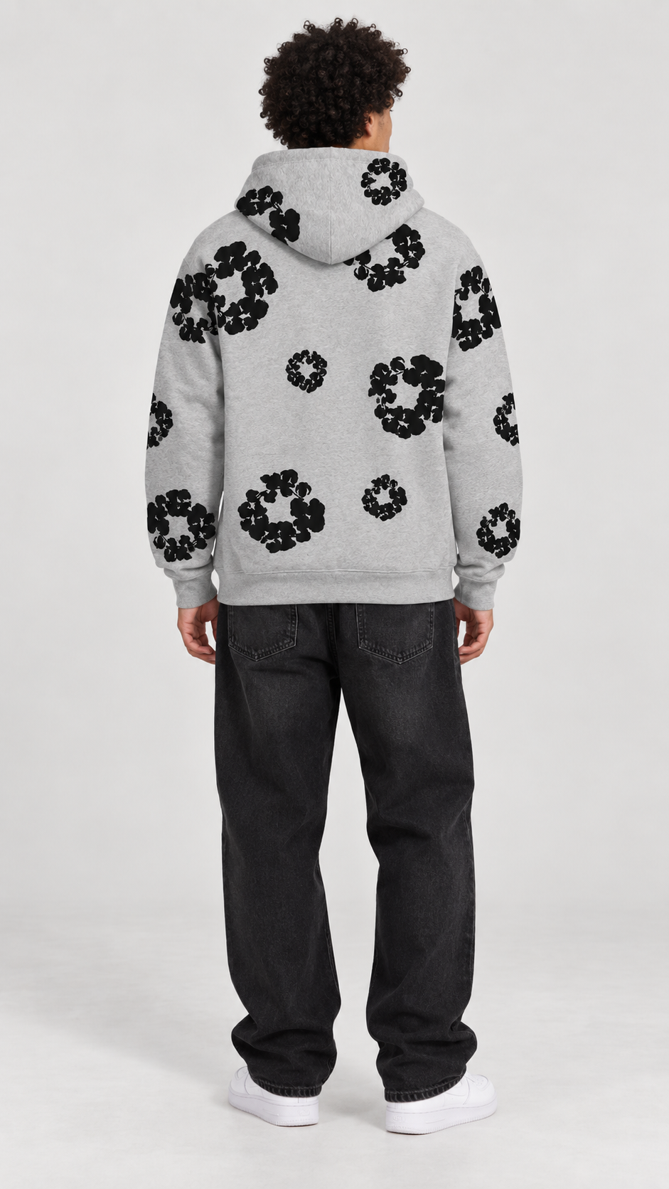 A model viewed from the back wearing a light gray hoodie with black circular floral patterns, paired with dark jeans and white sneakers, against a light gray studio background.
