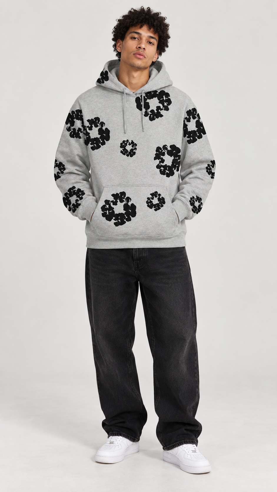 A man modeling a light gray pullover hoodie with black circular floral patterns on the front, paired with dark jeans and white sneakers, against a plain light gray studio background.