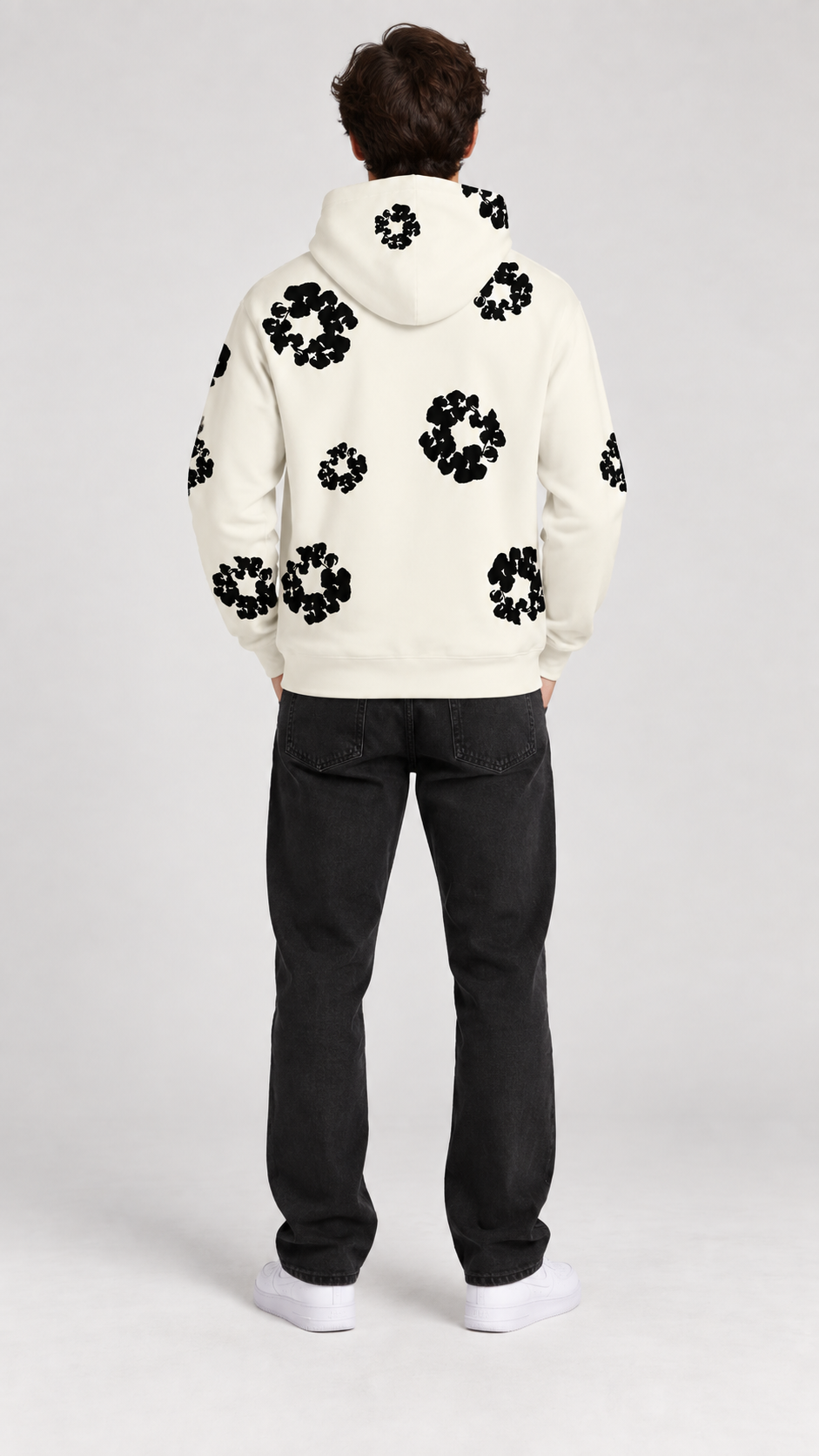 A man viewed from behind wearing a cream-colored hoodie with black floral patterns and black jeans, standing against a light gray studio background.