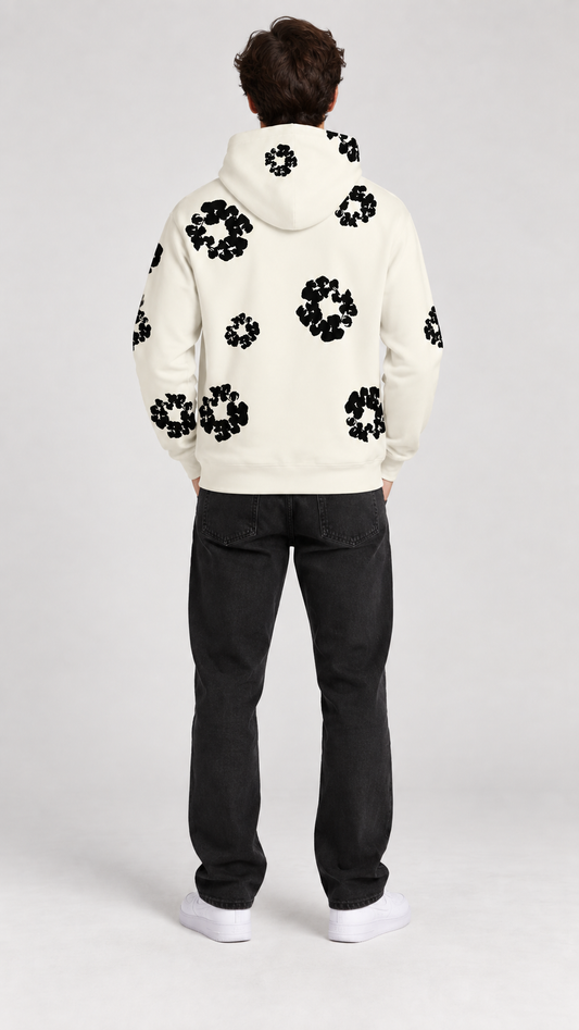 A man viewed from behind wearing a cream-colored hoodie with black floral patterns and black jeans, standing against a light gray studio background.