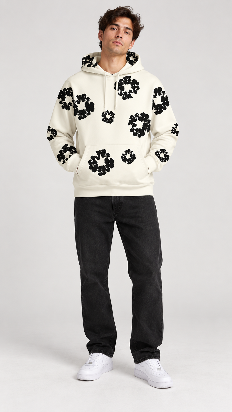 A man modeling a cream-colored hoodie with black circular floral patterns, paired with black pants and white sneakers, against a light gray studio background.