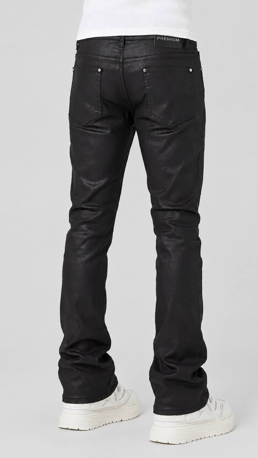 Black leather flare jeans shown from the back on a model wearing a white t-shirt and white platform sneakers, against a light gray studio background.