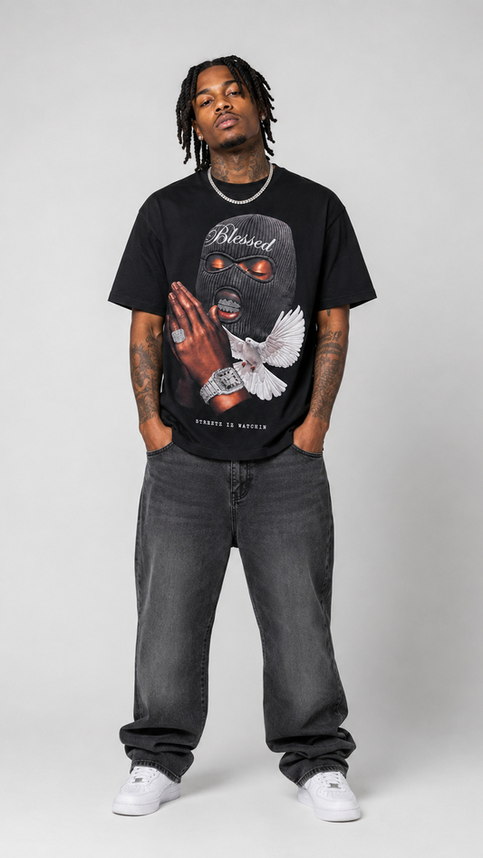 A man modeling a black graphic t-shirt with a 'Blessed' brand print, paired with dark gray jeans and white sneakers, standing against a light gray studio background.