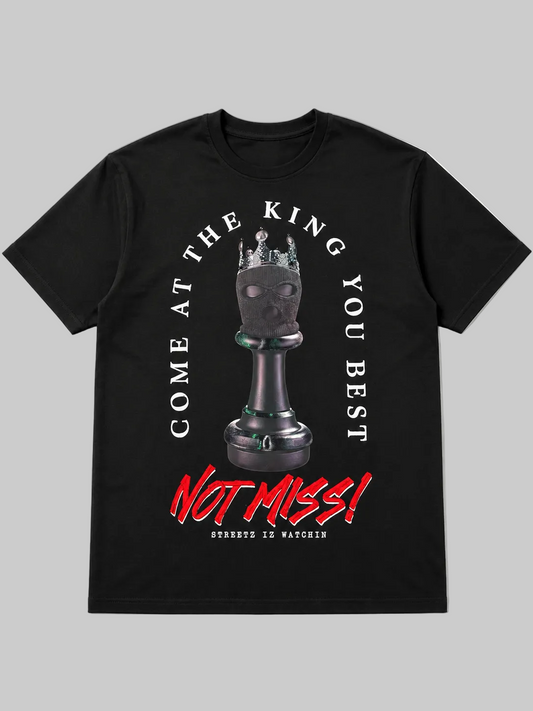 A black short-sleeve crewneck t-shirt featuring a graphic print of a chess king piece with a crown on its head. The text 'COME AT THE KING YOU BEST' is printed in white arching above the chess piece, and 'NOT MISS!' is printed in a bold red graffiti-style font below.