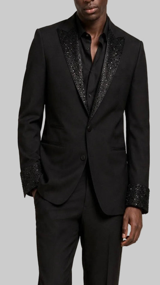 A man wearing a black tuxedo jacket with sparkling black sequined peak lapels and matching sequined cuff trim, paired with a black shirt and trousers, against a grey background.