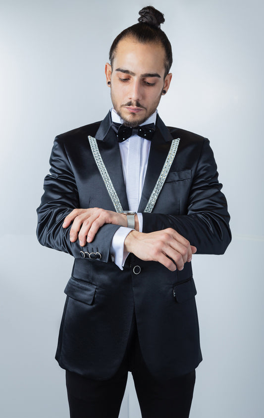 Black, one-button tuxedo jacket with a slim fit. The jacket features a shiny black fabric, rhinestone embellishments on the collar, a pocket square, and button cuffs. The jacket is made from a medium-lightweight fabric.
