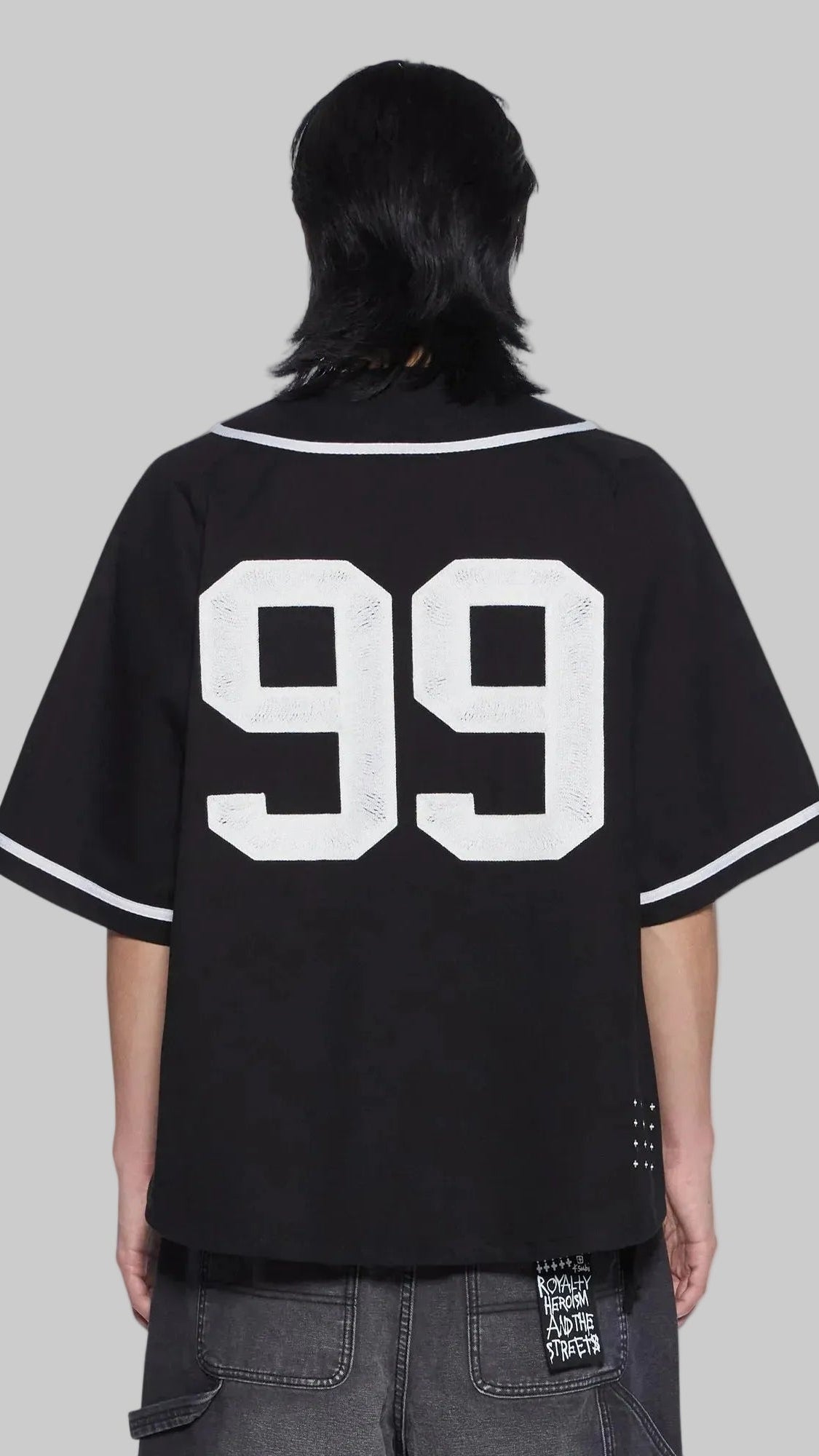 A person shown from the back wearing an oversized black baseball-style jersey with large white '99' lettering on the back and white trim on the collar and sleeve cuffs, paired with dark gray jeans against a plain white background.