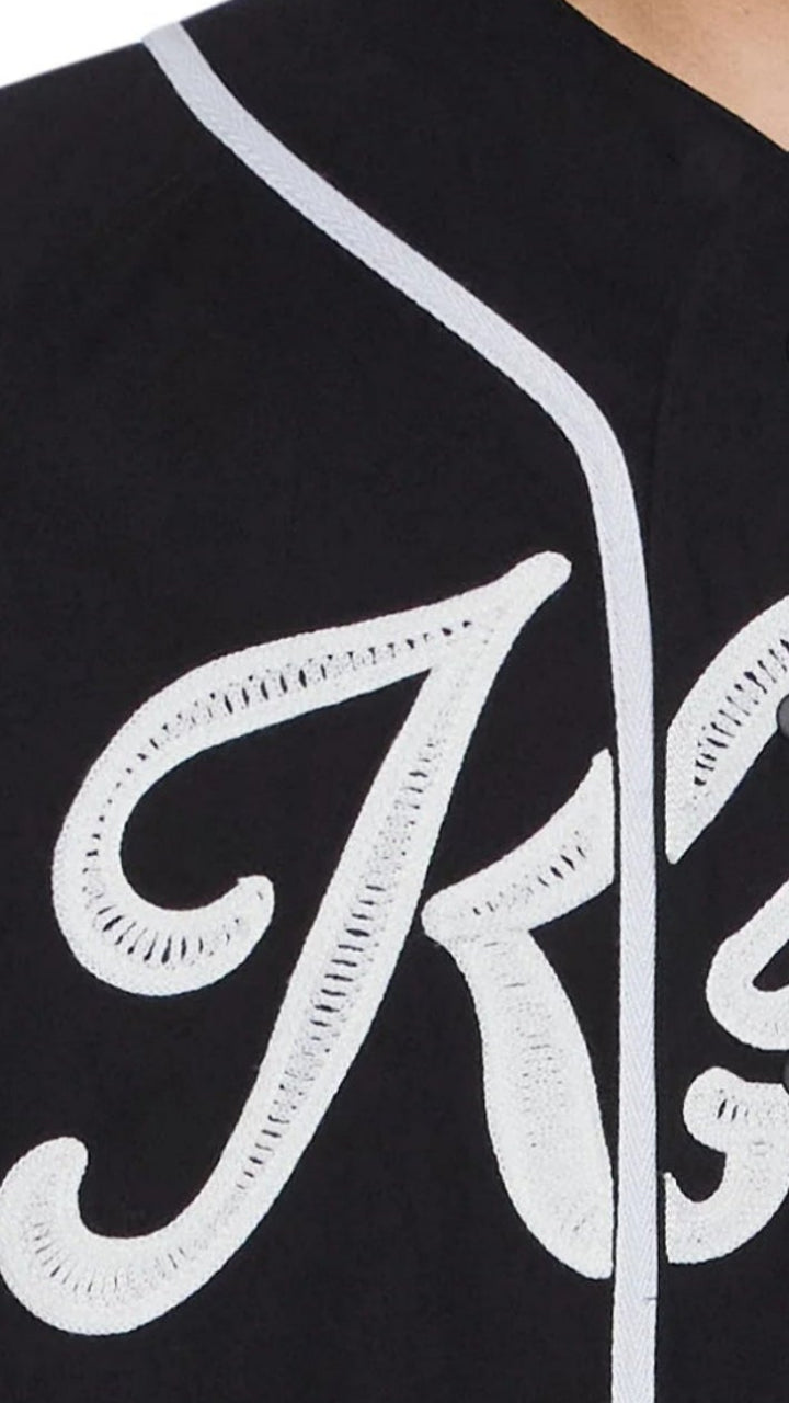 A close-up of a person wearing a black short-sleeve baseball-style jersey with white piping and a large white script logo across the front. The visible brand text reads 'Ksubi'.
