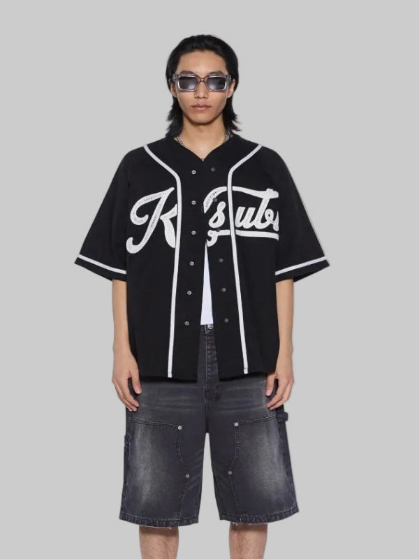A model stands against a white background wearing a black short-sleeve baseball-style shirt with white piping and a large white script logo across the chest, paired with dark gray denim shorts, white socks with red text, and black-and-white sneakers.