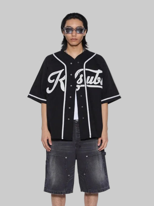 A model stands against a white background wearing a black short-sleeve baseball-style shirt with white piping and a large white script logo across the chest, paired with dark gray denim shorts, white socks with red text, and black-and-white sneakers.