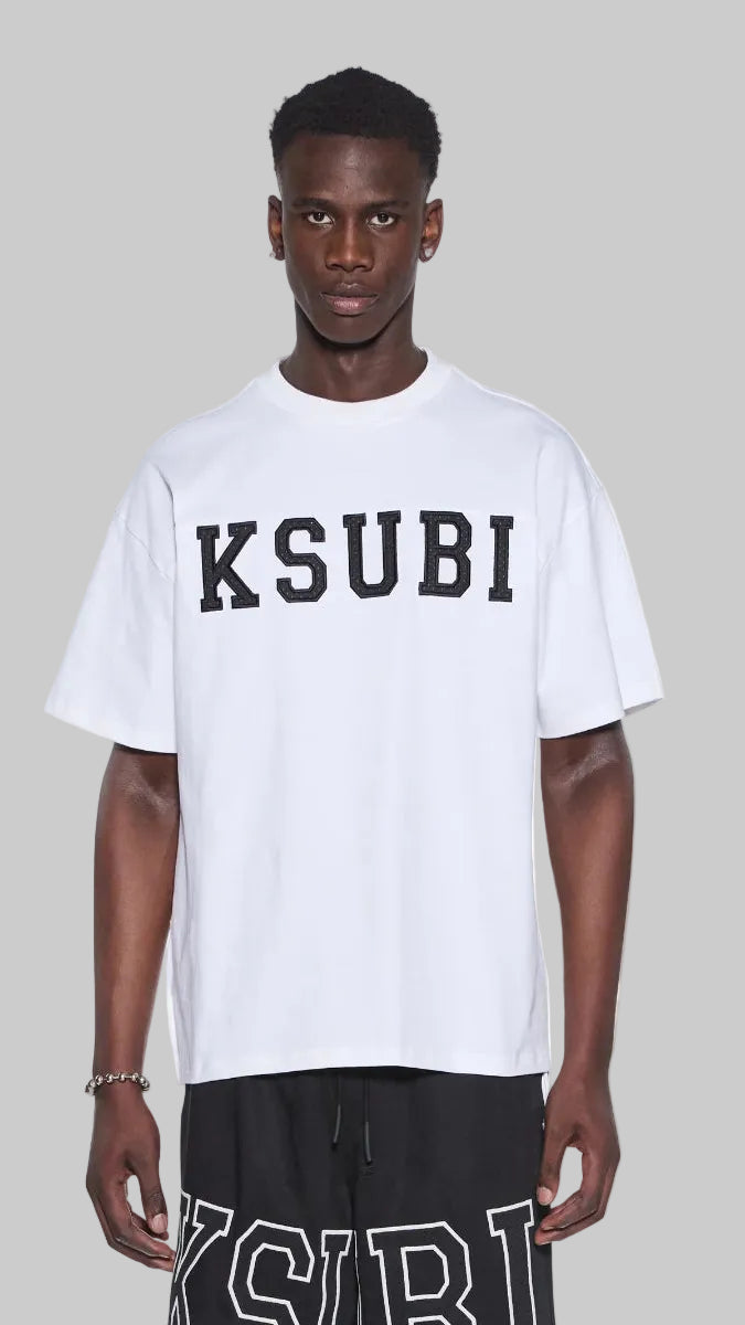 A man wearing a white oversized t-shirt with 'KSUBI' printed in black across the chest, paired with black shorts featuring white lettering, against a plain white background.