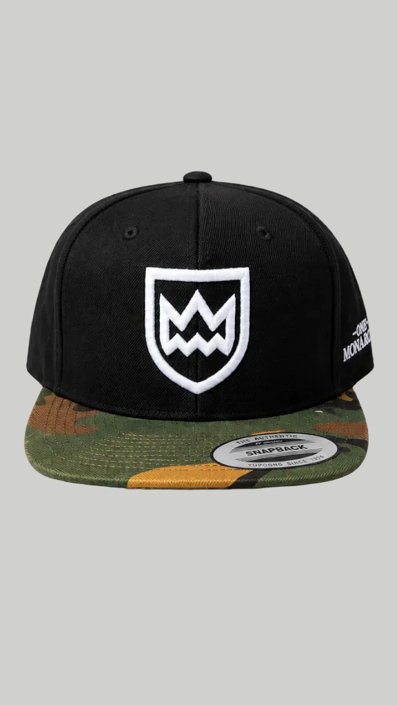 A black snapback cap with a white embroidered shield logo on the front and a green, brown, and tan camouflage flat brim, shown against a solid gray background.