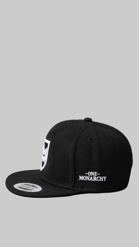 A black snapback cap featuring a white shield logo on the front panel and 'ONE MONARCHY' embroidered in white on the side, set against a solid gray background.