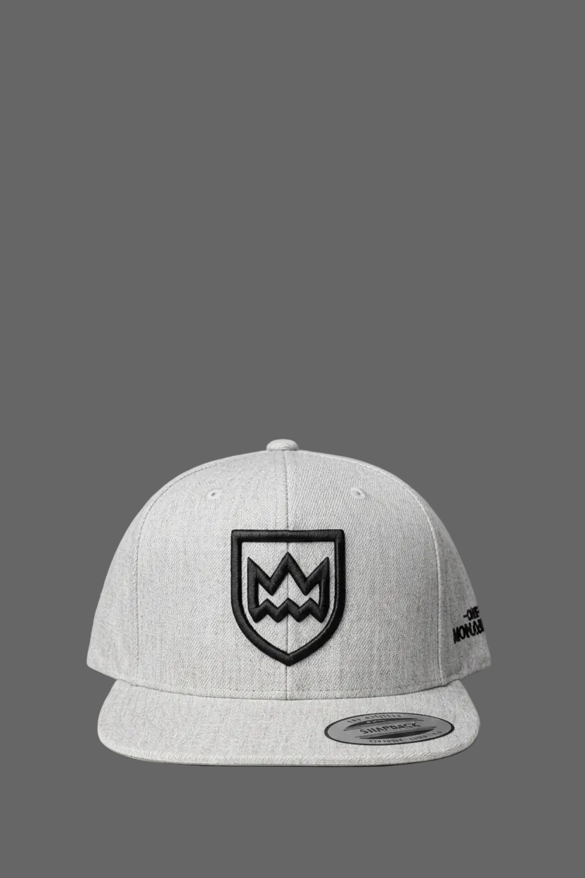 A light gray heather flat-brim cap featuring a black embroidered shield logo with a stylized 'M' on the front and a small black logo on the side, set against a solid dark gray background.
