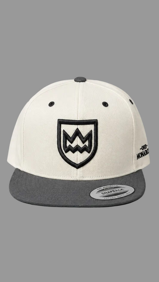 A structured snapback cap with an off-white crown and a dark gray flat brim. The front features a black embroidered shield logo with a stylized 'M' inside, and the side displays 'ONE MONARCHY' branding.