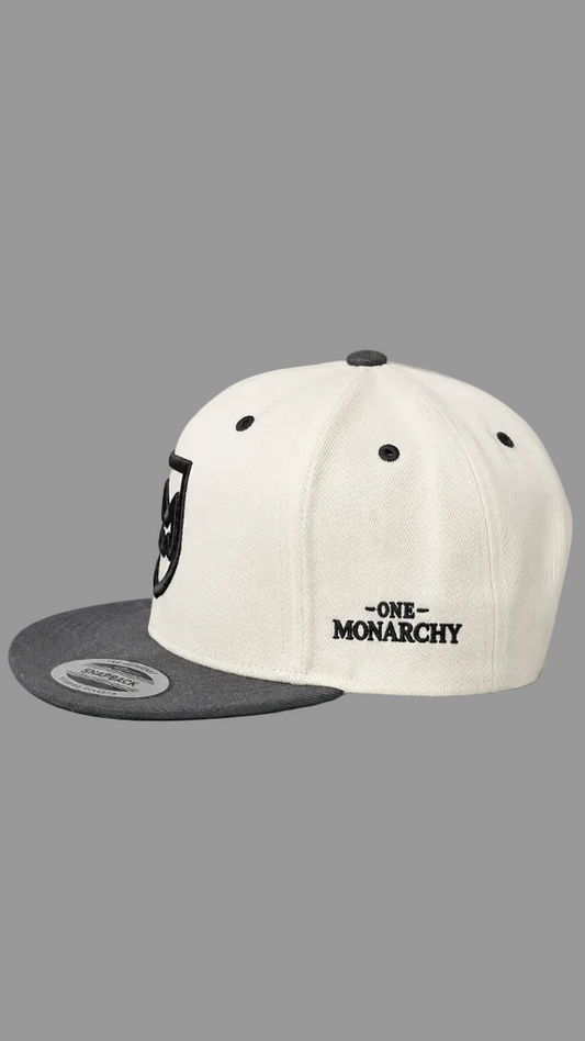 An off-white snapback cap with a dark gray flat brim, featuring the text '-ONE- MONARCHY' embroidered in black on the side. A black shield logo is visible on the front panel, and a silver metal sticker is on the brim.