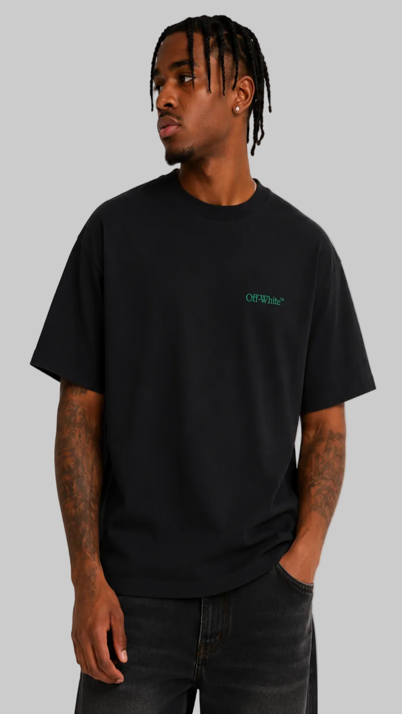 A man wearing a black crewneck t-shirt with a small green 'OFF-WALL' logo on the left chest, paired with dark jeans, photographed against a light gray studio background.