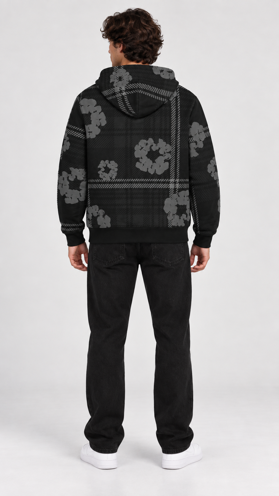 A man viewed from the back wearing a black hooded sweatshirt with a gray plaid pattern and gray floral motifs, paired with black pants and white sneakers, against a light gray studio background.
