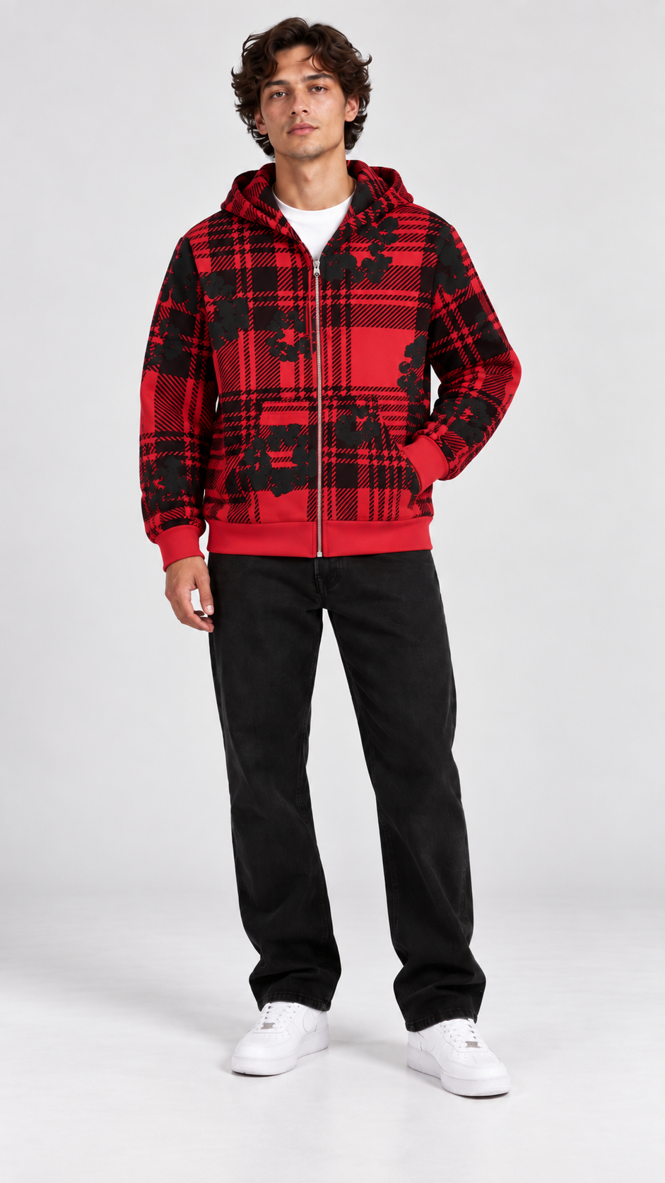 A man modeling a red and black plaid zip-up hoodie with a black graphic print, paired with black sweatpants and white sneakers, against a light gray studio background.