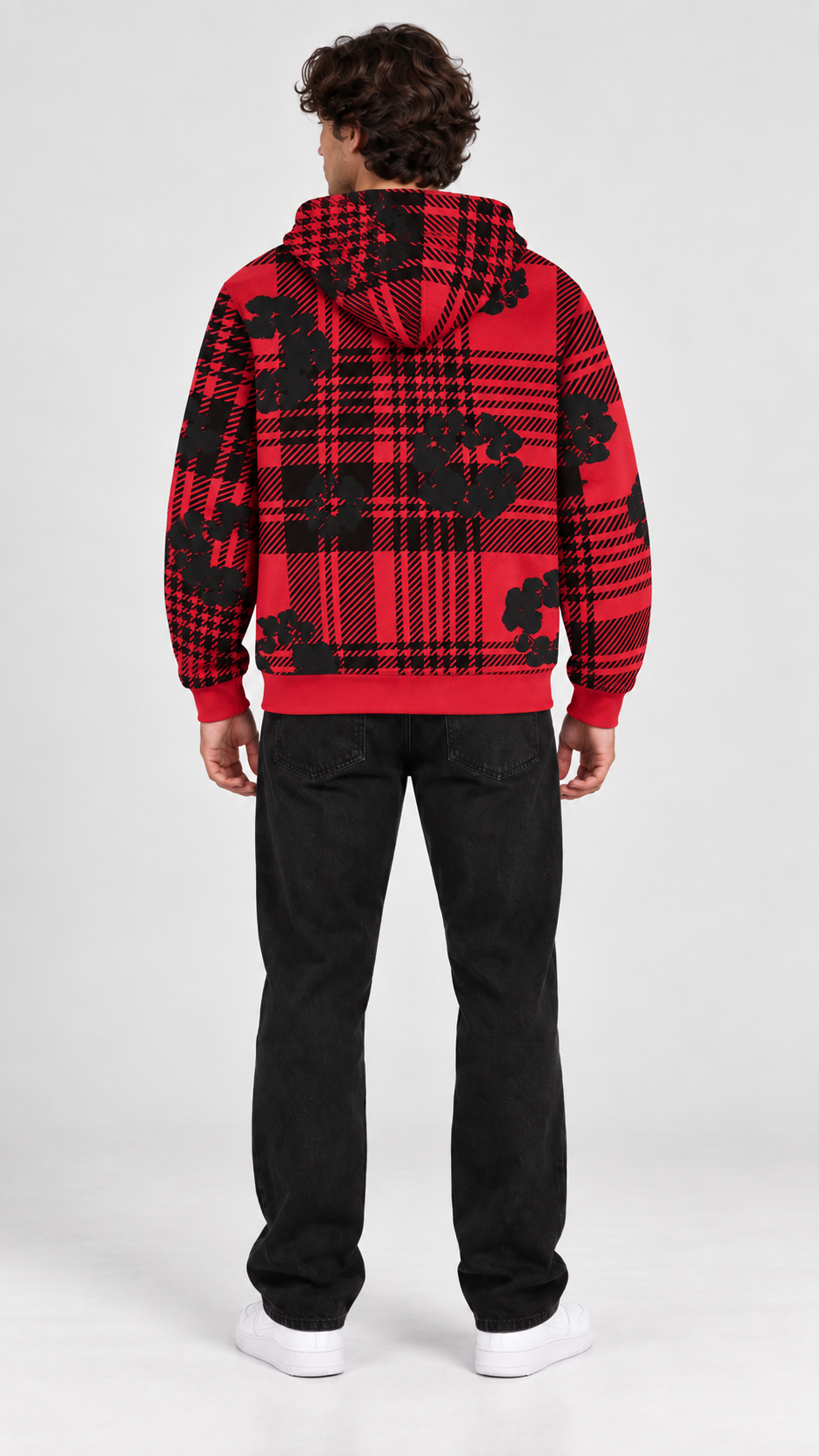 A man viewed from the back wearing a red and black plaid hoodie with black floral motifs, paired with black jeans and white sneakers, against a light gray studio background.