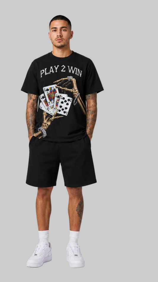 A male model shown from the front wearing a black short-sleeve graphic T-shirt with 'PLAY 2 WIN' text and playing cards printed on the front, paired with black shorts, white socks, and white sneakers against a light gray studio background.