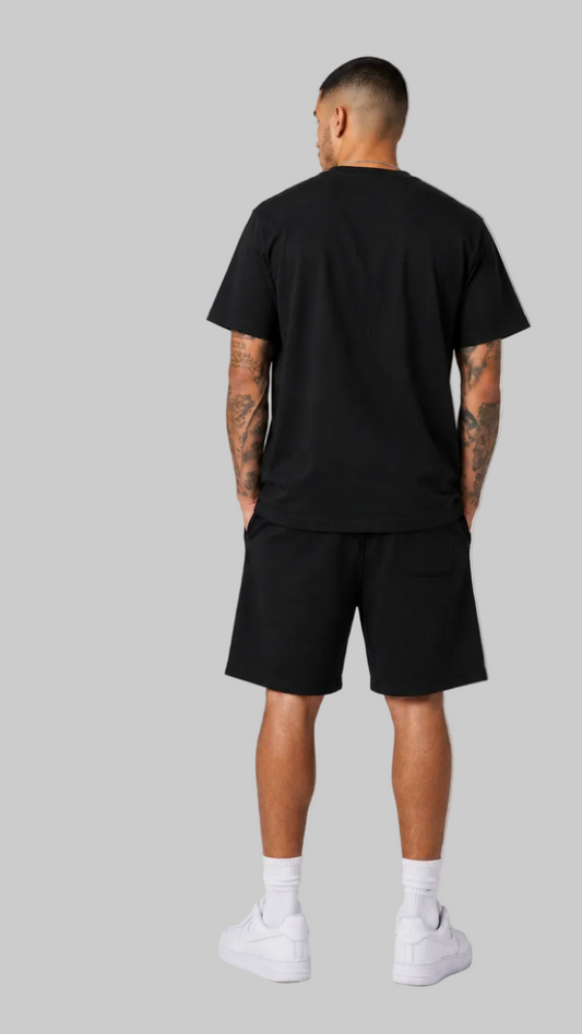 A male model shown from the back wearing a black short-sleeve graphic T-shirt paired with black shorts, white socks, and white sneakers against a light gray studio background.