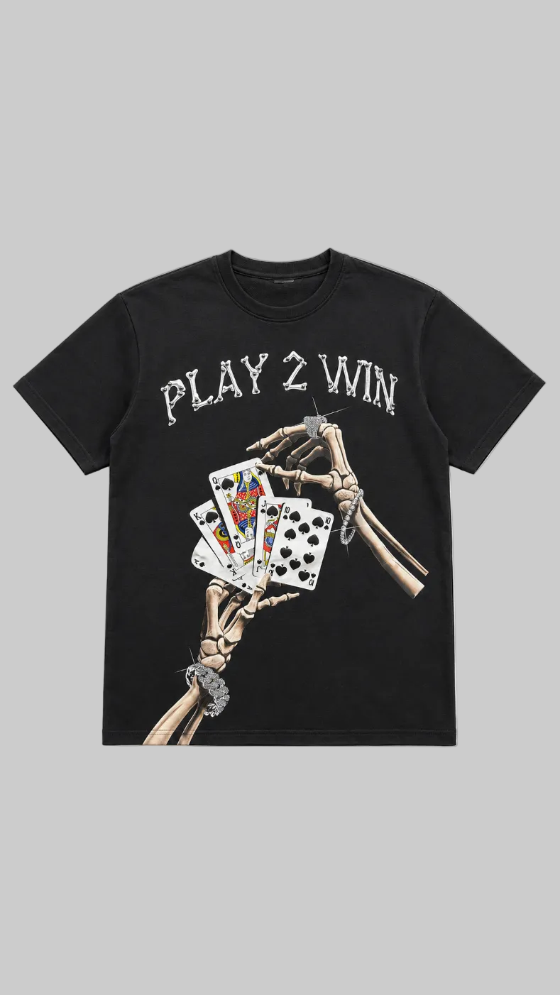A black short-sleeve graphic t-shirt featuring the text 'PLAY 2 WIN' in a distressed white font above a graphic of skeletal hands holding playing cards, displayed on a light gray background.