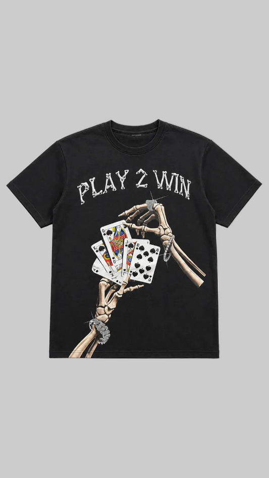 A black short-sleeve graphic t-shirt featuring the text 'PLAY 2 WIN' in a distressed white font above a graphic of skeletal hands holding playing cards, displayed on a light gray background.