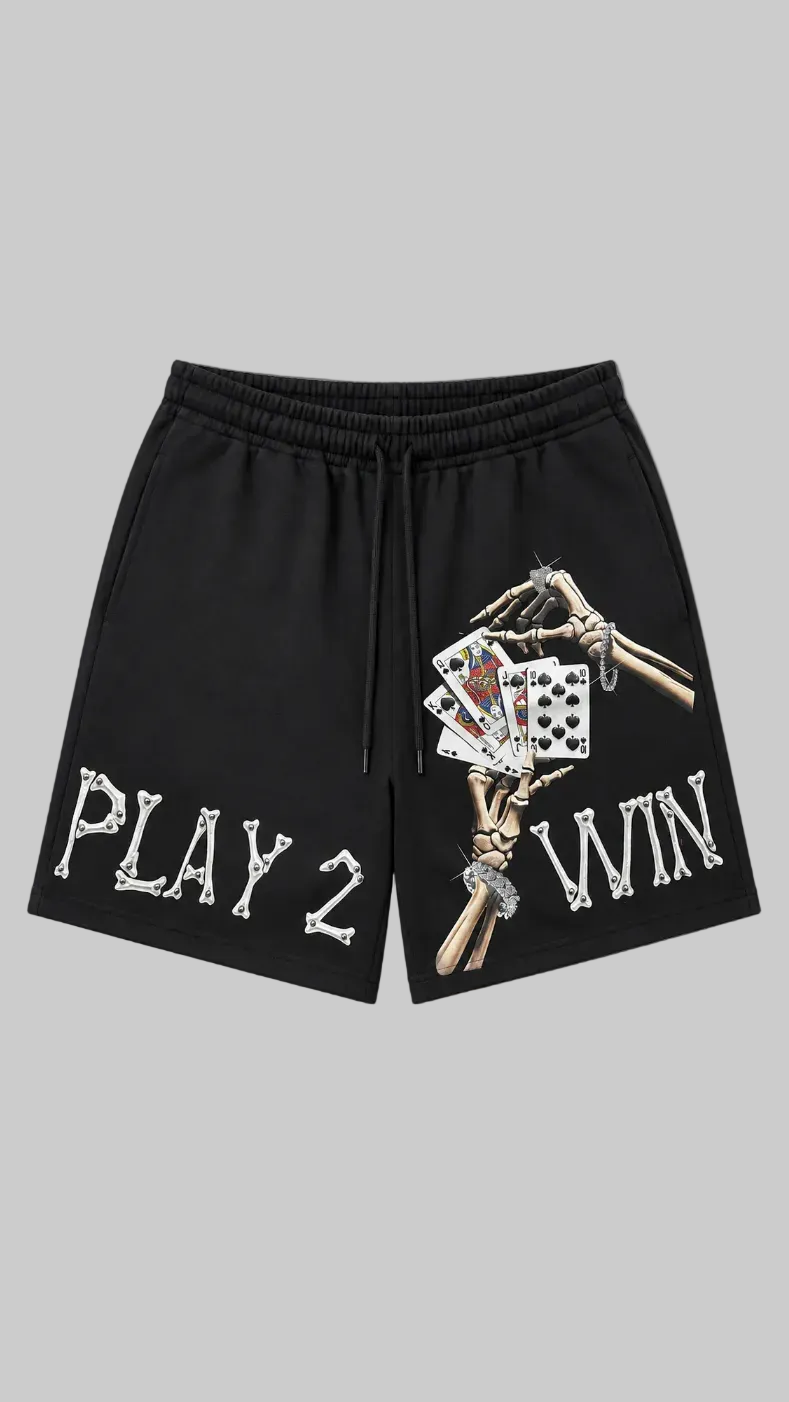 Black athletic shorts with a graphic print of skeleton hands holding playing cards on the right leg, and the text 'PLAY 2 WIN' in a bone-style font on the left leg, shown against a light gray background.