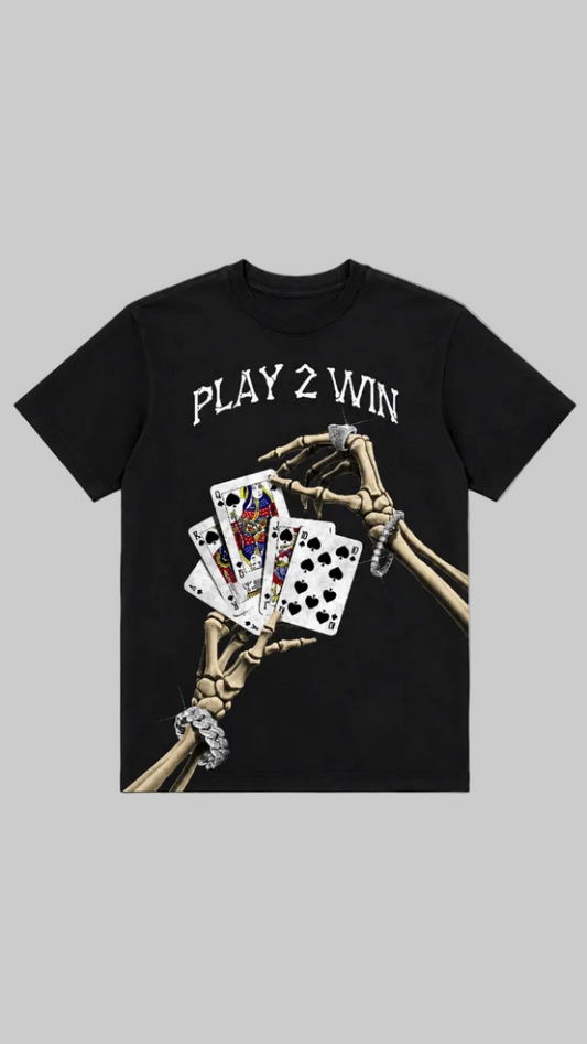 A black short-sleeve graphic t-shirt featuring the text 'PLAY 2 WIN' in white above a graphic of skeletal hands holding playing cards, one of which has a diamond ring, against a light gray background.