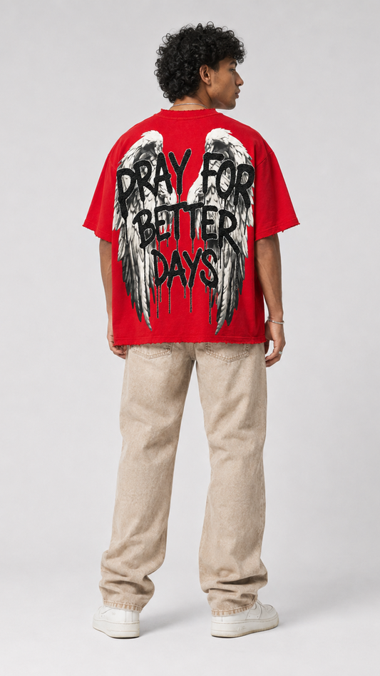 A man shown from the back wearing a bright red graphic t-shirt with a large black and white angel wings design and the text 'PRAY FOR BETTER DAYS' printed across the back, paired with beige pants and white sneakers, against a light gray studio background.