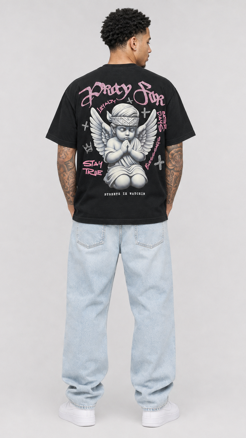 A man viewed from behind wearing a black graphic t-shirt with a large cherub print on the back, paired with light-wash jeans and white sneakers, against a light gray studio background.