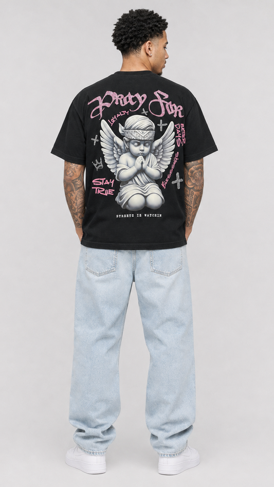 A man viewed from behind wearing a black graphic t-shirt with a large cherub print on the back, paired with light-wash jeans and white sneakers, against a light gray studio background.